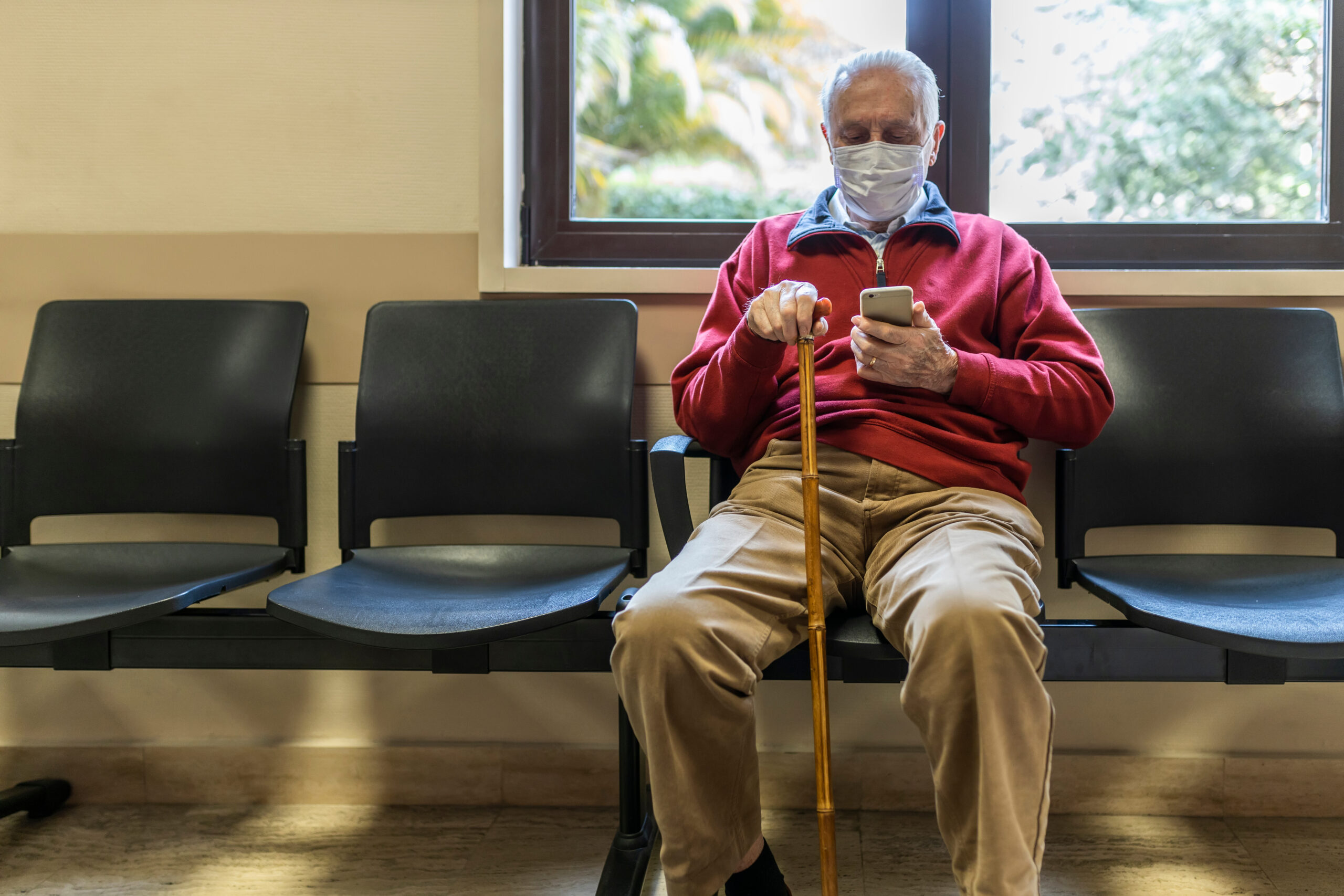 Elderly man holding a cane sitting at the doctor’s office with a mask on.