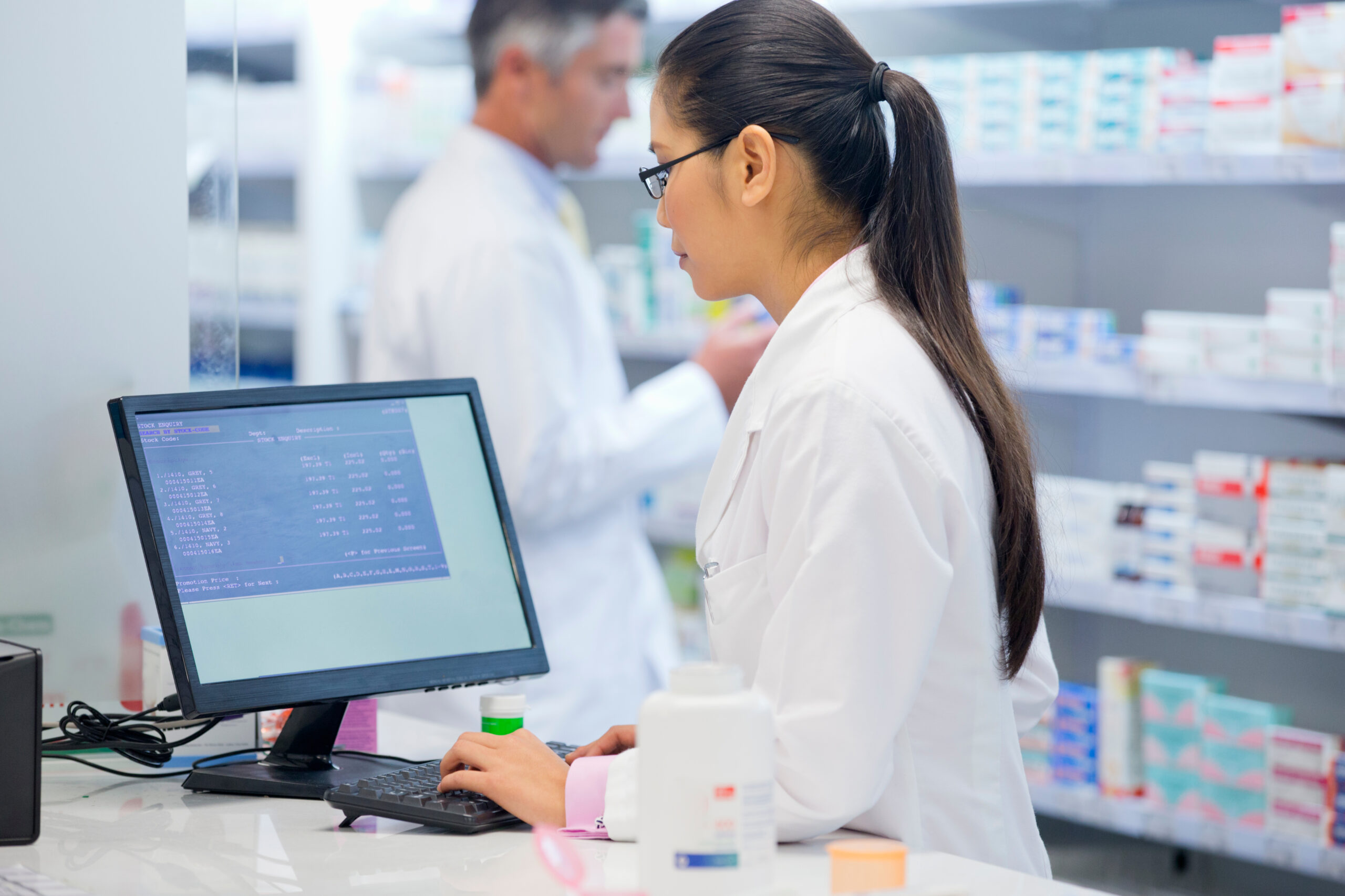 A female pharmacist working on a computer with a male pharmacist in the background.