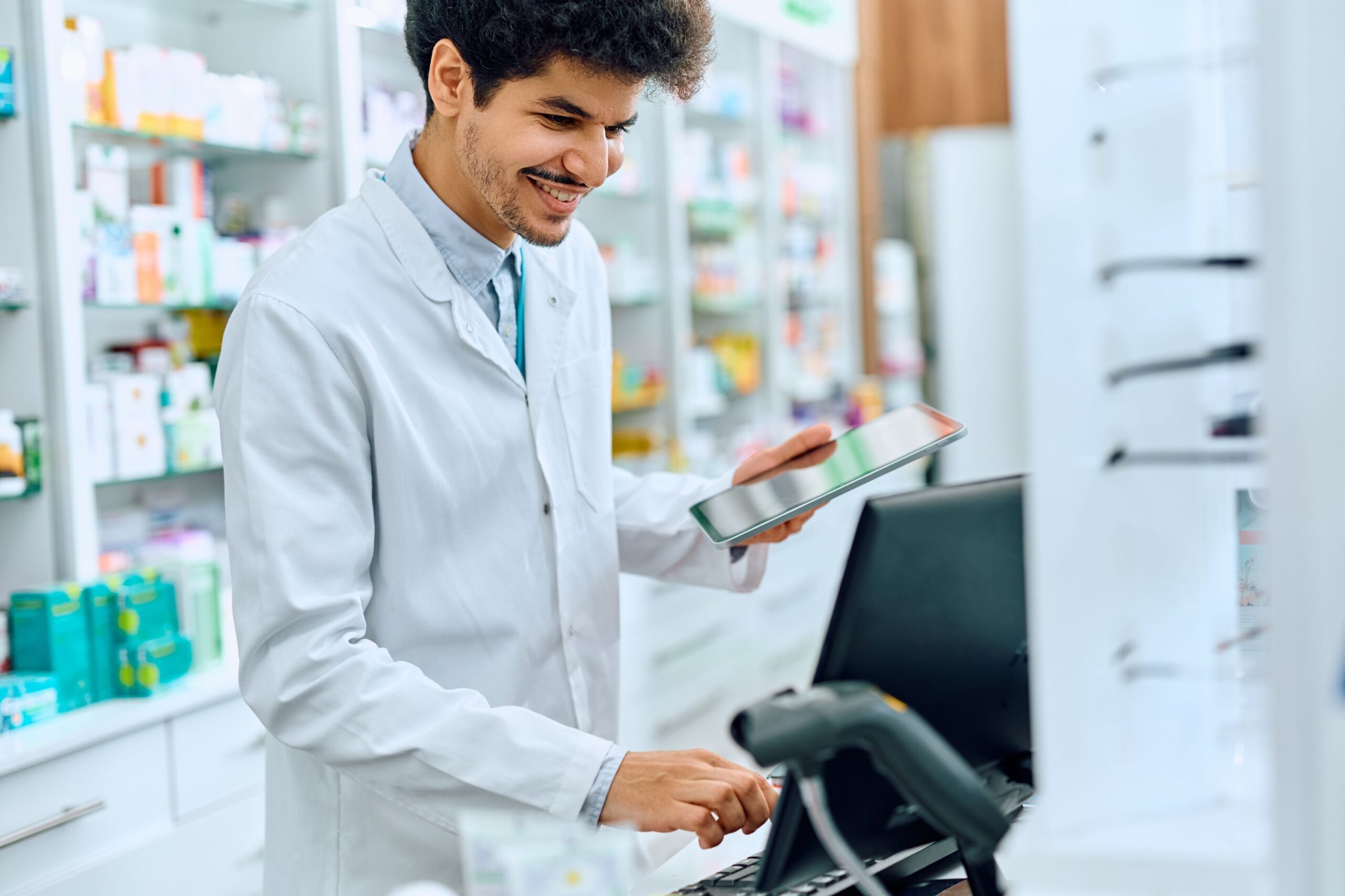 A male pharmacist typing on a computer while holding a tablet.