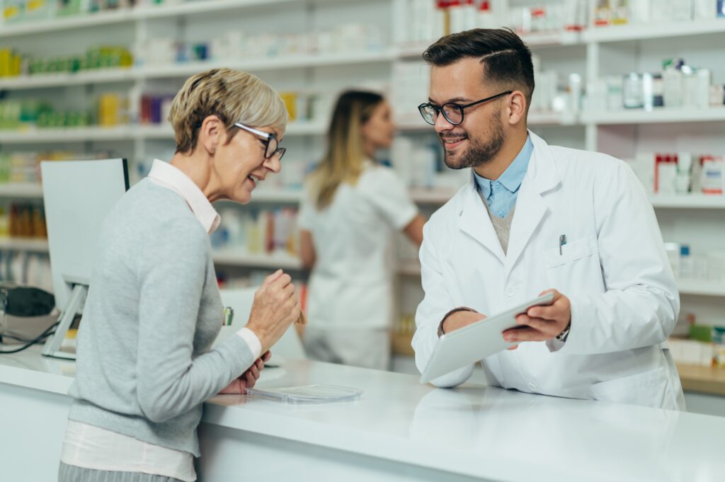 A male pharmacist using a tablet to help a elderly female customer.