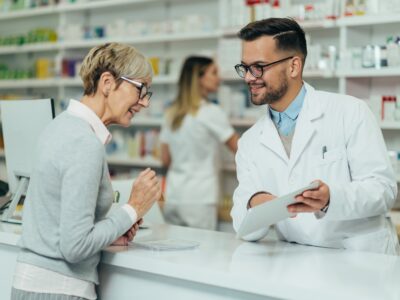 A male pharmacist using a tablet to help a elderly female customer.