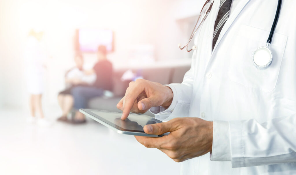 A male doctor tapping on a tablet with patients in the waiting room in the background.