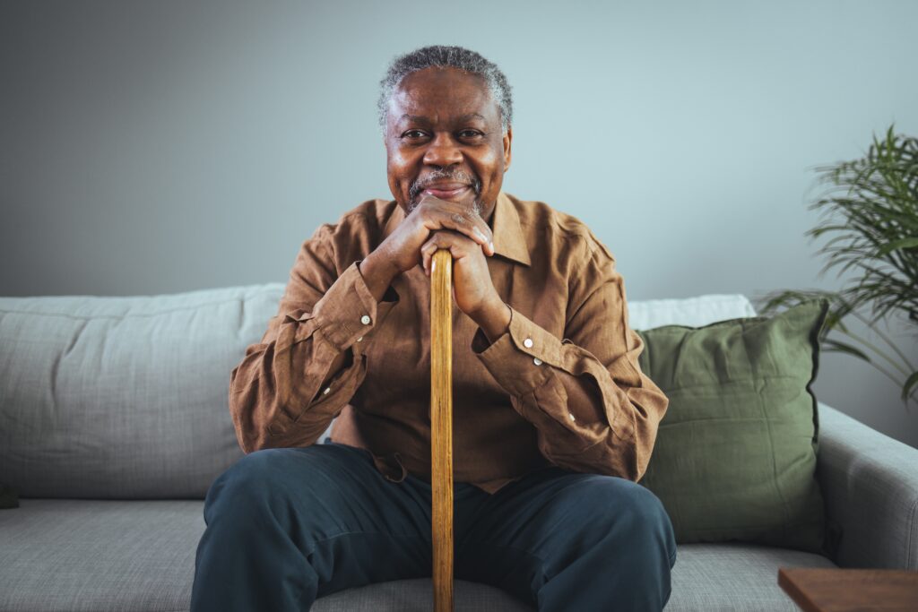 A smiling elderly man sitting on couch while holding his cane.