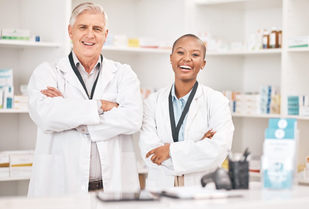 A male and female pharmacist with crossed arms smiling at the pharmacy.