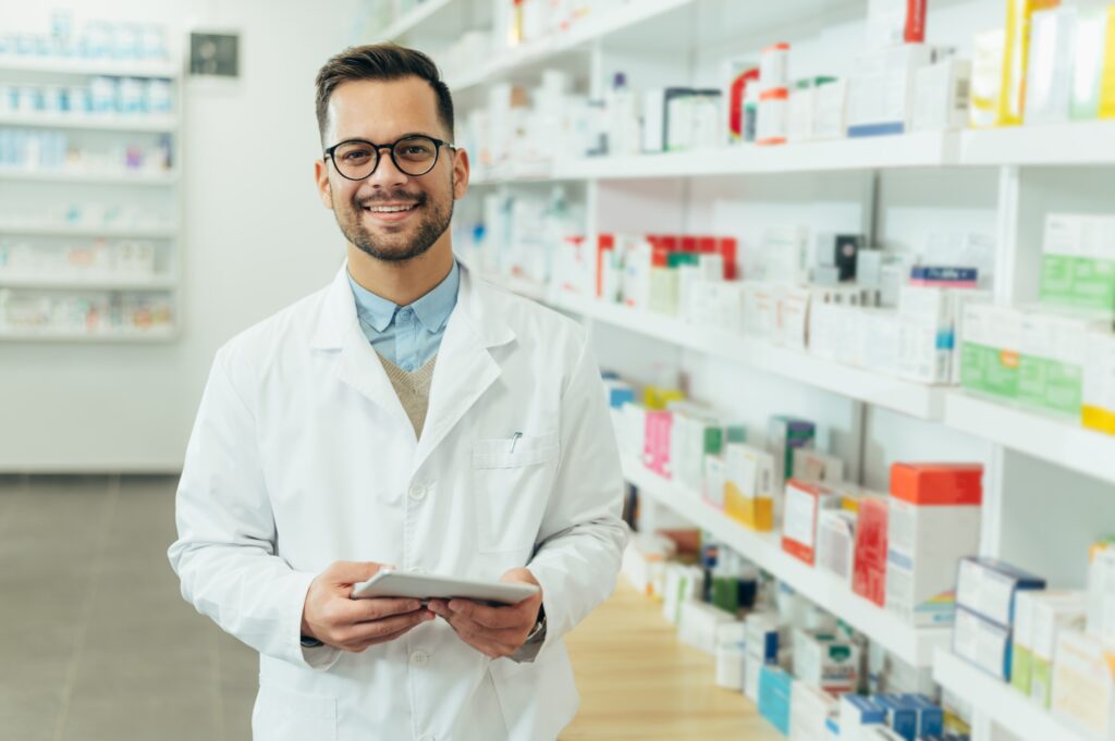 Male pharmacist holding a tablet with the pharmacy in the background.
