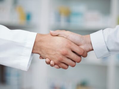 A doctor and a pharmacist shaking hands in front of a blurred background.